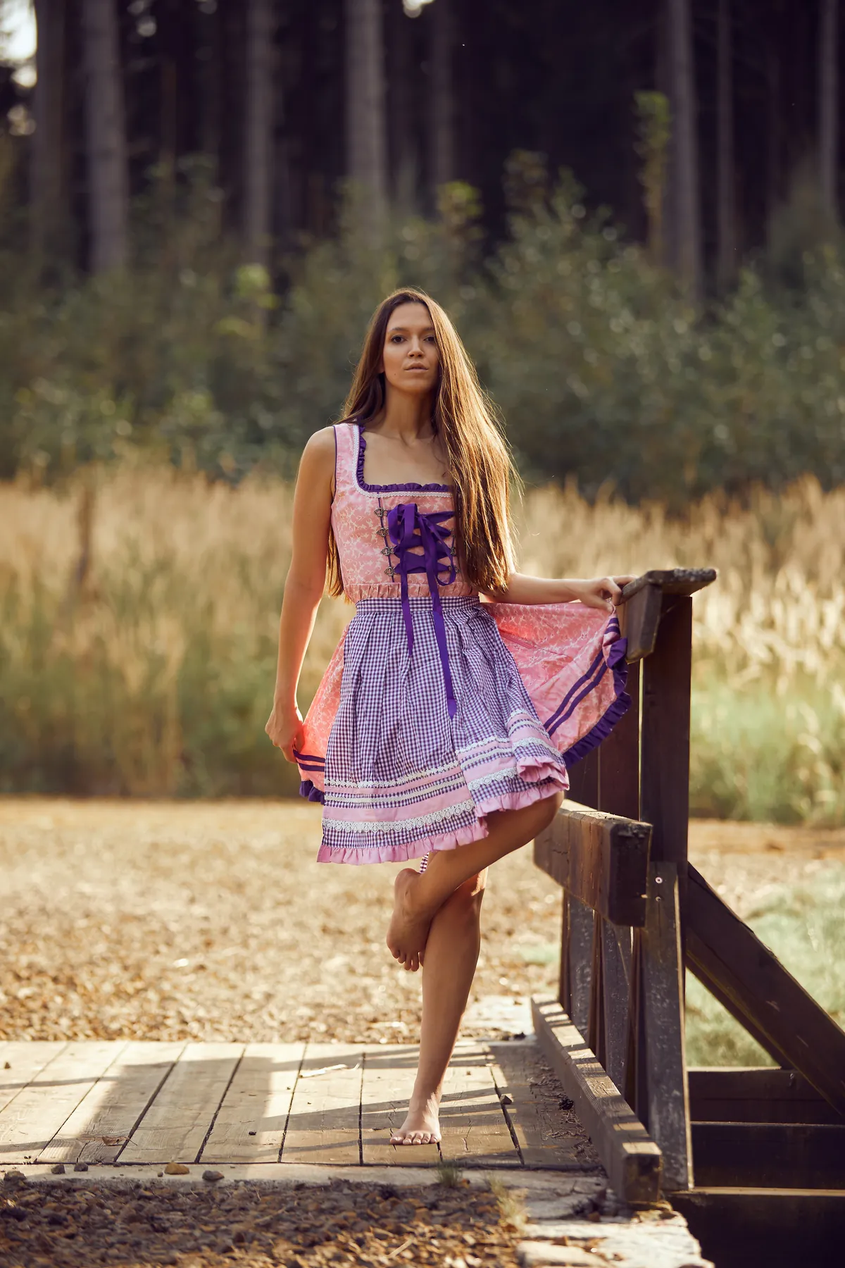 Model im lila Dirndl steht barfuß auf einem Holzsteg am Waldrand