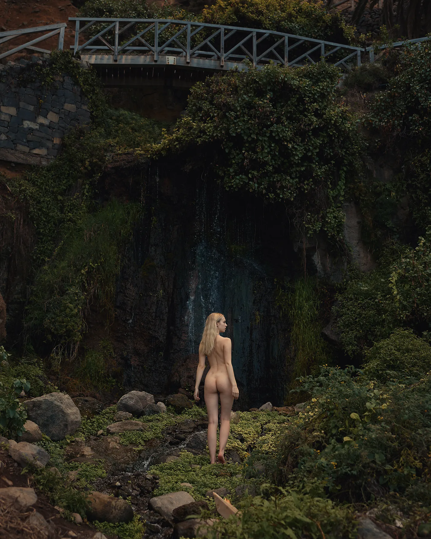 Model from behind in front of an overgrown waterfall with a small bridge above it