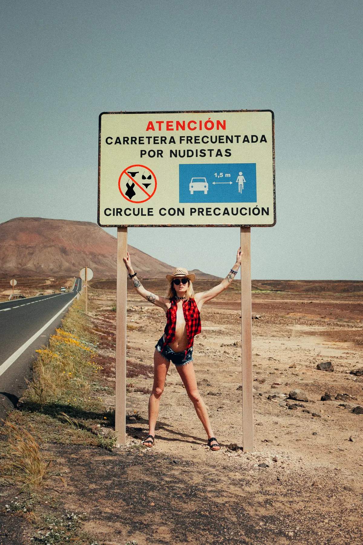 Model with a straw hat and open checked shirt below a road sign on a country road in a barren landscape